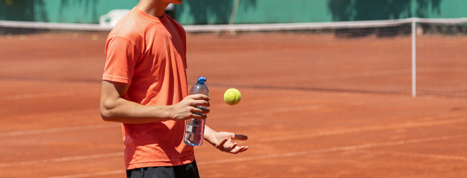 Teenage tennis player holding water bottle while tossing tennis ball during hydration break on an outdoor clay court in sunny weather. Wide banner with copy space for text or design