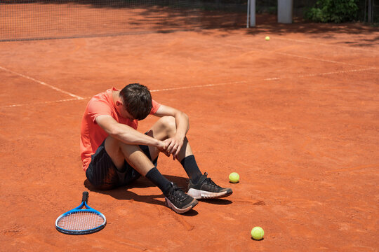 Teenage tennis player sitting on clay court after losing match, expressing disappointment, frustration and emotional exhaustion during competition with copy space.