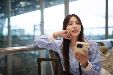 Obraz premium Young Asian Woman Waiting at Airport Terminal Holding Smartphone with Luggage