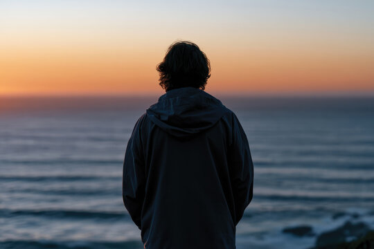 A person stands in contemplation, gazing out at the ocean as the sun sets, creating a poignant moment that evokes feelings of reflection and serenity.