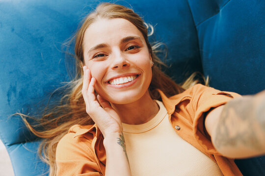 Close up young woman wear brown shirt do selfie shot pov on mobile cell phone lay down blue sofa stay at home hotel flat rest relax spend free spare time in living room indoors. People lounge concept.