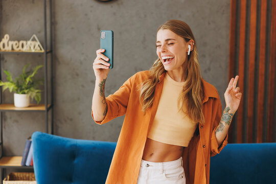 Young fun cool woman wear brown shirt listen music in earphones use mobile cell phone dancing near blue sofa stay at home flat relax spend free spare time in living room indoor. People lounge concept