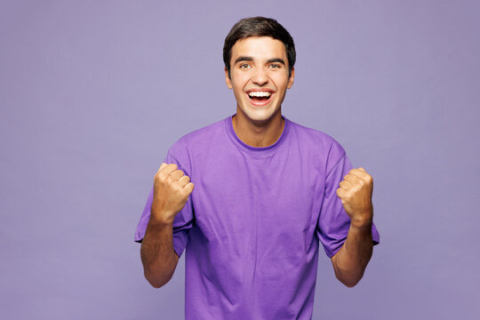 Young Caucasian man he wears violet t-shirt casual clothes doing winner gesture celebrate clenching fists say yes isolated on plain pastel light purple background studio portrait. Lifestyle concept.