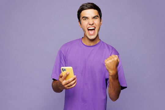Young happy Caucasian man he wear violet t-shirt casual clothes hold in hand use mobile cell phone do winner gesture isolated on plain pastel light purple background studio portrait Lifestyle concept
