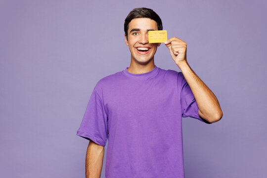 Young Caucasian man he wear violet t-shirt casual clothes hold in hand cover eye with mock up of credit bank card isolated on plain pastel light purple background studio portrait. Lifestyle concept.