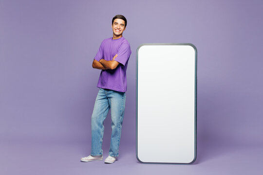 Full body young man he wear violet t-shirt casual clothes stand near big huge blank screen mobile cell phone smartphone with workspace copy space mockup area isolated on plain light purple background.