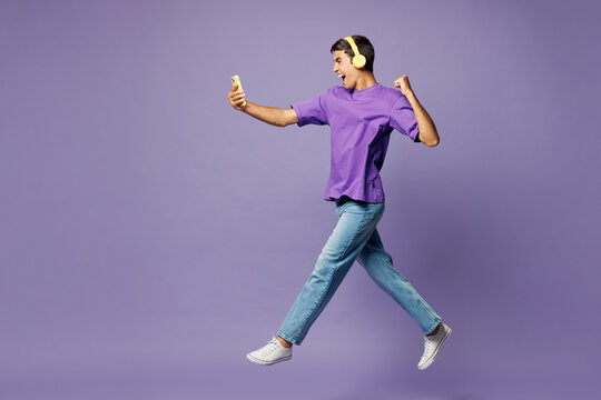 Full body young man he wear violet t-shirt casual clothes listen to music in headphones hold use mobile cell phone do winner gesture sing isolated on plain pastel purple background Lifestyle concept.