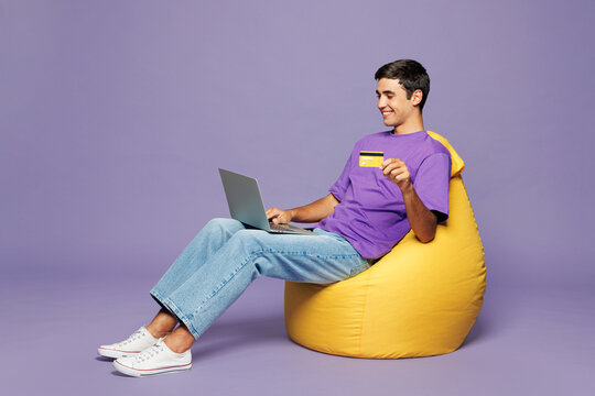 Full body young IT man wear violet t-shirt casual clothes sit in bag chair using laptop pc computer hold credit bank card shopping online order delivery book tour isolated on plain purple background.