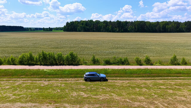 Car parked along a rural dirt road beside vast green fields under a bright blue sky, aerial view