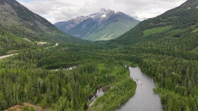 Aerial drone footage of a pristine mountain valley with a reflective pond dense pine forest a winding road and snowcapped peaks in the distance along the Canadian Rockies in Revelstoke, BC.