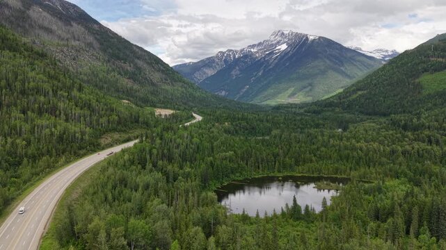 Aerial drone footage of a pristine mountain valley with a reflective pond dense pine forest a winding road and snowcapped peaks in the distance along the Canadian Rockies in Revelstoke, BC.