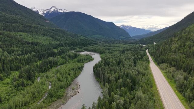 Aerial drone footage of a pristine mountain valley with a reflective pond dense pine forest a winding road and snowcapped peaks in the distance along the Canadian Rockies in Revelstoke, BC.