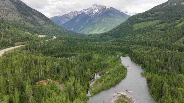 Aerial drone footage of a pristine mountain valley with a reflective pond dense pine forest a winding road and snowcapped peaks in the distance along the Canadian Rockies in Revelstoke, BC.