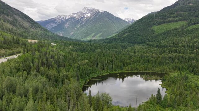 Aerial drone footage of a pristine mountain valley with a reflective pond dense pine forest a winding road and snowcapped peaks in the distance along the Canadian Rockies in Revelstoke, BC.