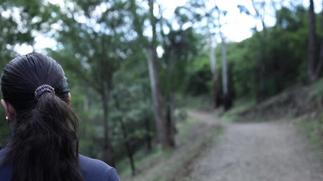 A woman with dark hair in a ponytail walks a winding dirt trail through lush forest at Mt Lofty, near Melbourne. Soft daylight, steady camera, tranquil atmosphere, natural sounds.