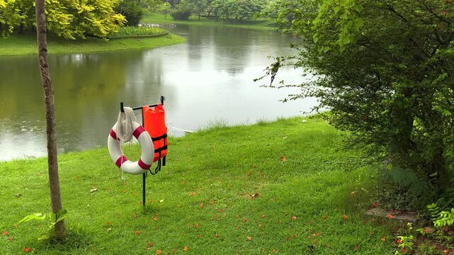 A tranquil, static shot of a serene public park during a gentle rain. In the foreground, a red and white lifebuoy ring, an orange life jacket, and a coiled rope hang on a metal post.