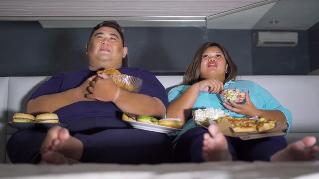 Overweight Asian couple sitting on couch at night watching television while eating junk food including pizza, burgers, fried chicken, donuts, and popcorn.