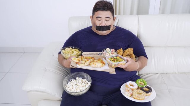 Overweight Asian man sitting on couch with black adhesive tape on his mouth trying to stop eating a variety of junk foods like pizza, donuts, and fried chicken.