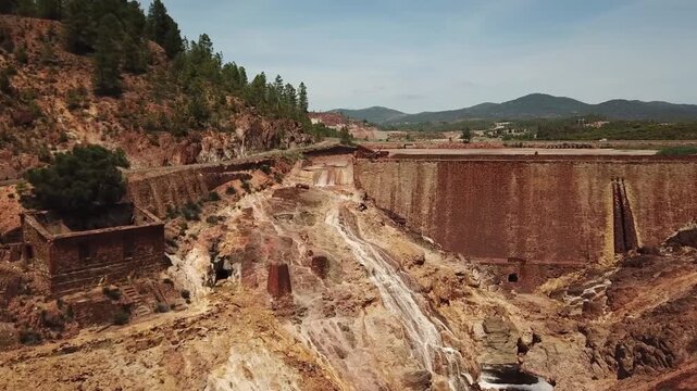 short video clip capturing a small, mineral-rich waterfall flowing over a historic dam wall at the Rio Tinto river. In the distant background, a lonely road cuts through the rugged landscape