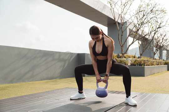 Fit woman doing kettlebell sumo squat exercise on yoga mat outdoors, wearing black sportswear and smartwatch. Strength training, lower body workout, healthy lifestyle and fitness concept.