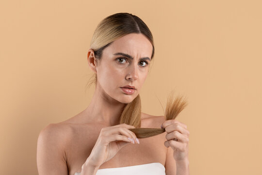 Concerned young woman examining damaged split ends of her hair, showing frustration with haircare problem on beige studio background, closeup