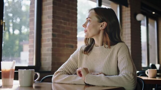Scrolling woman in cream sweater holding smartphone at cafe, pausing and gazing outside with mug
