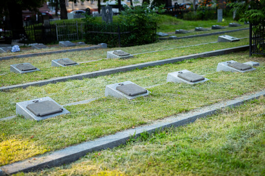 Rows of flat grave markers on grassy cemetery grounds with soft sunlight, trimmed hedges, stone pathways, and calm memorial setting across maintained lawn, with grave, marker, cemetery, memorial