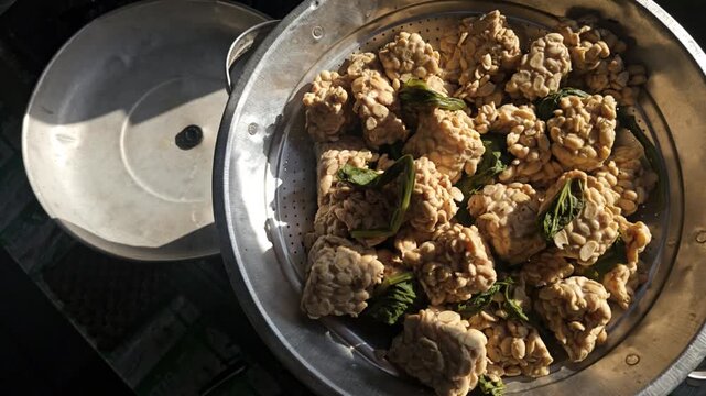 Raw tempeh chunks with green leaves in a metal steamer pot ready for cooking under natural sunlight