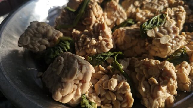 Macro view of fresh uncooked tempeh slices derived from Glycine max with green leaves resting in a metal pan ready for healthy cooking