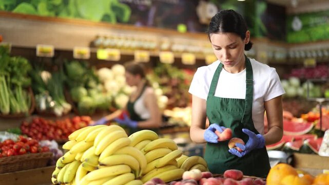 Adult woman seller in apron puts fresh peaches on display in vegetable shop