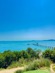 Fototapeta premium Bright sunny day with a view of a long pier stretching into the blue sea and trees lining the shore at a coastal location