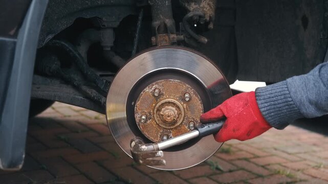 Gloved hands striking seized brake disc with hammer for removal from wheel hub. Automotive repair loosening stuck brake rotor during car maintenance with manual force. Closeup dismantling corroded