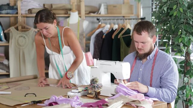 Focused male tailor sewing pastel fabric at machine with precision, while young female colleague sketching patterns on kraft paper nearby in shared studio