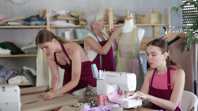 Team of tailors at work, three women in dressmaker office workroom. Seamstress staff sew, perform edge finishing stitching seam, assistant work with mannequin, designer draw dress pattern on paper