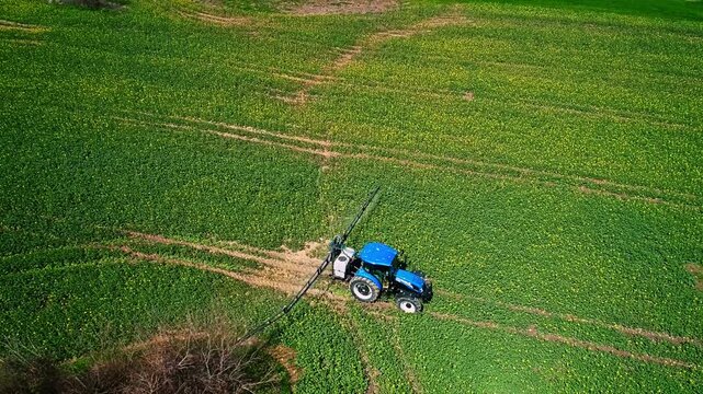 A drone view shows a tractor-mounted sprayer working on green farmland, performing agricultural spraying for crop protection, field maintenance, and modern farming practice.