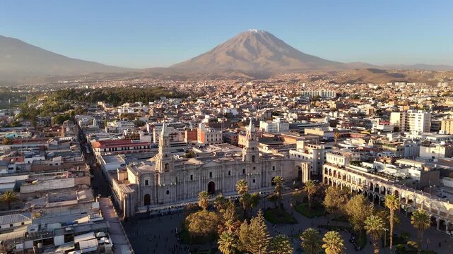 An aerial flybackward shot pulls away from the Basilica Cathedral to reveal the bustling Plaza de Armas in Arequipa, Peru, with El Misti volcano behind