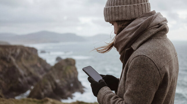 Woman smartphone traveler checks device on windy coastal cliff in cold weather showing navigation connection safety update during seaside trip  remote travel communication concept abroad