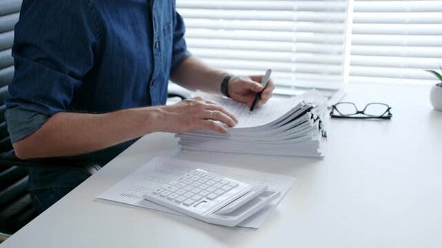 Individual working diligently on financial documents, calculating expenses and reviewing reports at an office desk, managing a large stack of paperwork and invoices