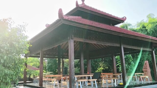 Traditional Javanese wooden gazebo or pendopo pavilion in a lush green garden resort, outdoor restaurant setup with wooden tables and chairs under a classic terracotta tile roof.