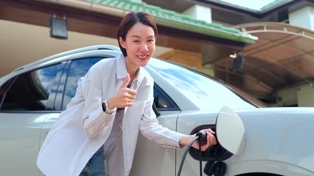 Smiling Asian woman showing thumbs up while plugging charger cable into an electric car at home. Concept of eco-friendly transport satisfaction and smart energy.