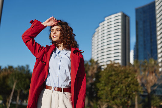 Young woman with curly hair wearing red coat and white shirt standing outdoors in city park with modern buildings in background on sunny day.