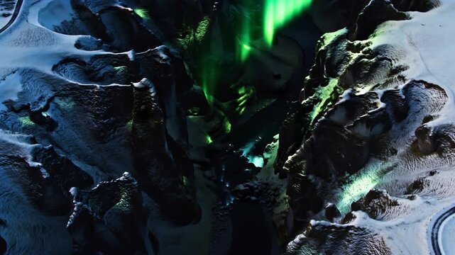 Streaming green auroral curtains descending over deep rocky canyon, lighting snow ridges and road