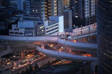 夕暮れ時の都市の夜景。高層ビル群と複雑な立体交差する道路、車のヘッドライトやテールライトが光る。 © BRUCE