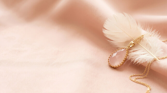 Rose quartz teardrop pendant on a gold chain resting on draped pink silk fabric with a white feather. Elegant jewelry flat lay with copy space