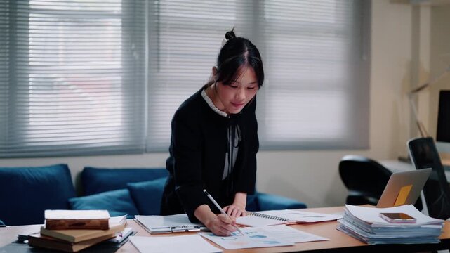 Young Asian Businesswoman Reviewing Documents at Office Desk with Laptop and Paperwork