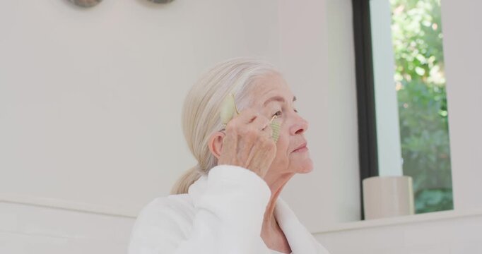 Senior female in white robe picking green facial tool, massaging face upward at vanity for skincare