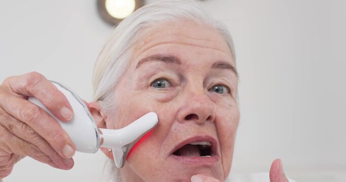 Senior woman touching skin with tri-tip lighting red LED moving left-right cheeks by sink for care