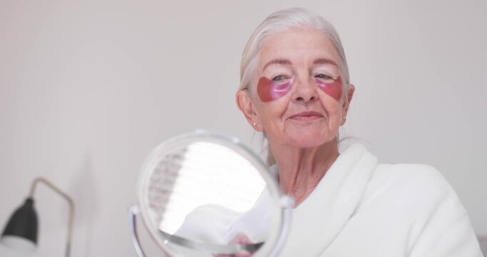 With pink heart patches Senior woman sitting at vanity in robe holding mirror adjusting patches