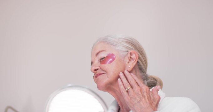 Senior female in robe starting skincare at vanity, applying pink eye patches while checking mirror