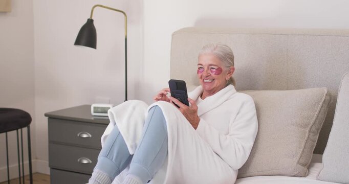 Senior woman sitting on bed tapping smartphone, reading, wearing white robe and pink eye patches